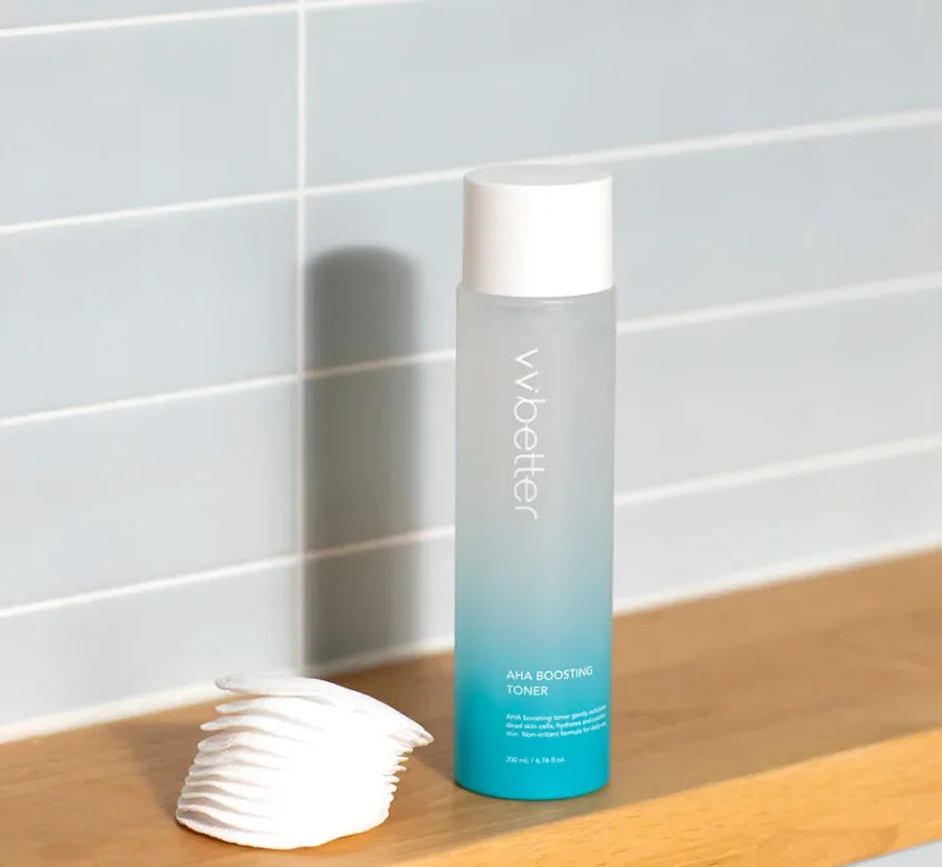 AHA Boosting Toner bottle on a wooden shelf beside a stack of cotton pads in front of a grey tiled wall