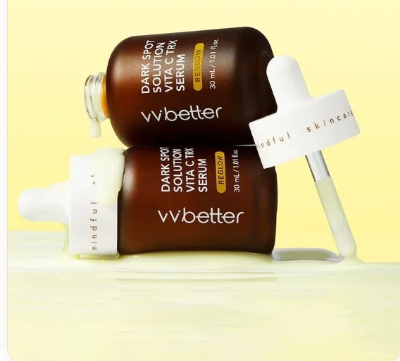 Two VVBETTER Dark Spot Solution Vita C TRX Serum bottles lying on a pale yellow serum pool, with the dropper cap resting against them