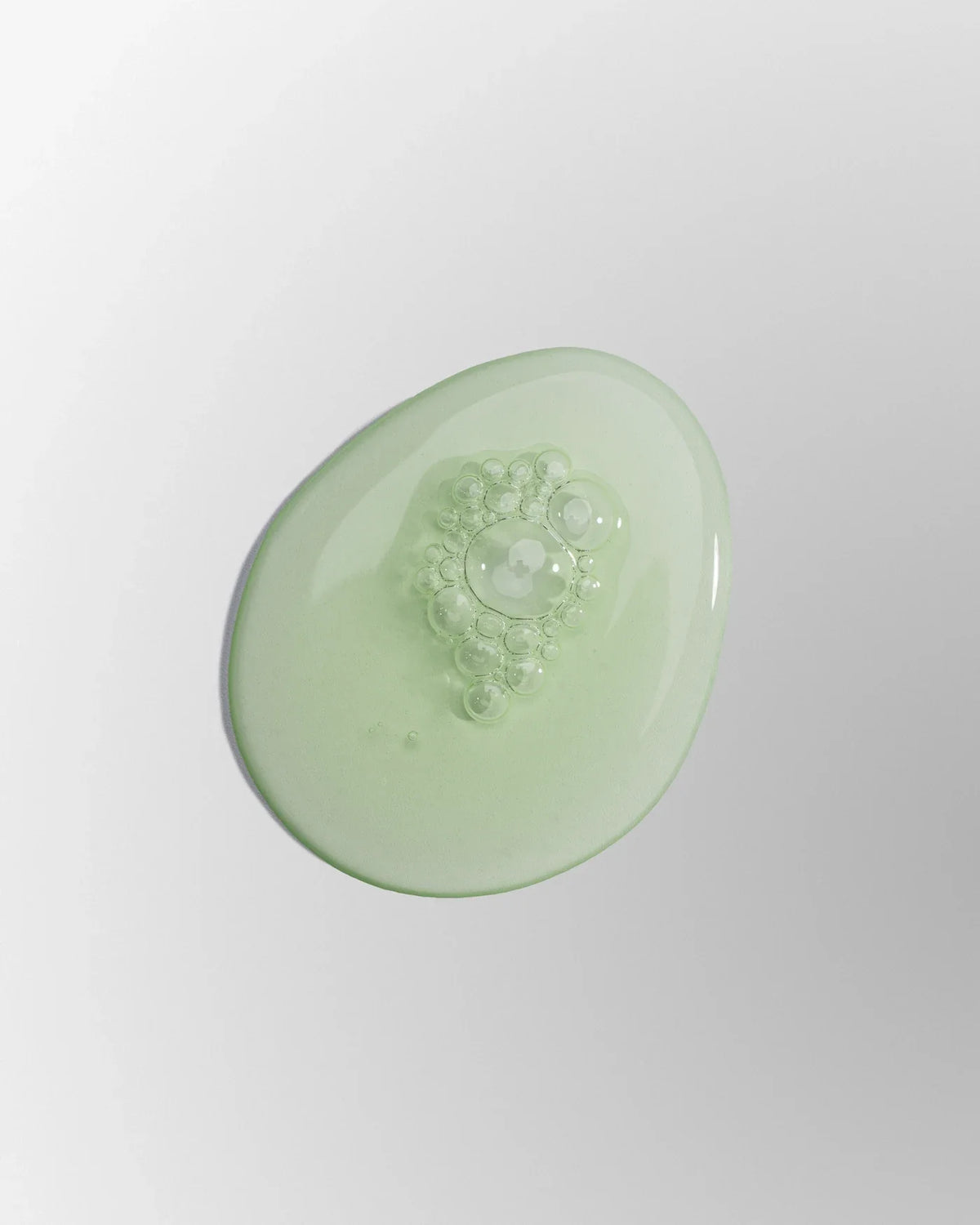 A clear green gel swatch with small bubbles on a light grey surface, from the Jeju Yuja Succinic Balancing Serum editorial photograph