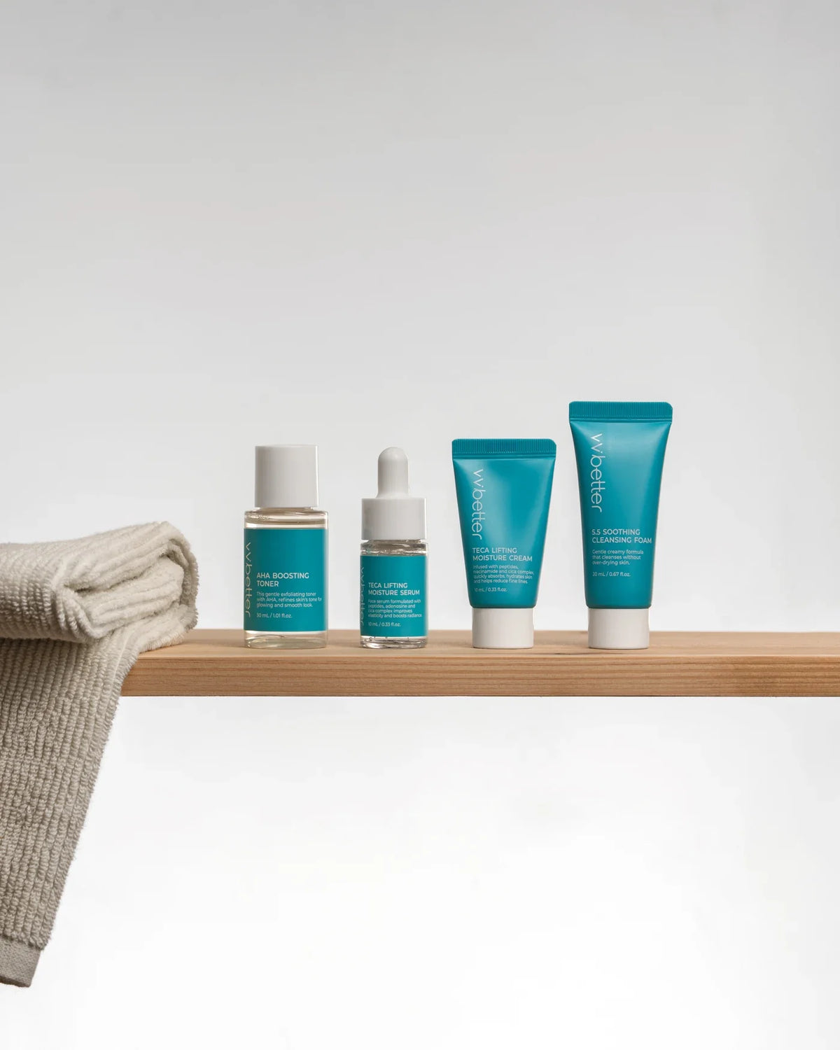 Four small VVBETTER Blue Line travel-size bottles and tubes standing on a wooden shelf beside a folded towel