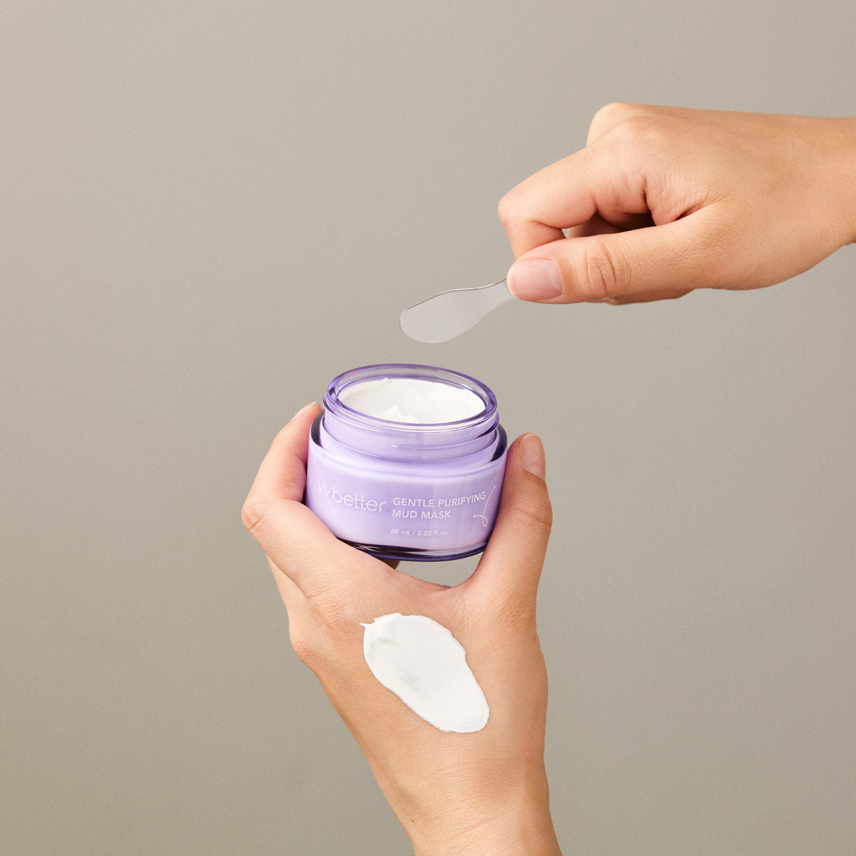 Gentle Purifying Mud Mask — open jar held in a hand with a spatula lifting the clay, swatch on the hand